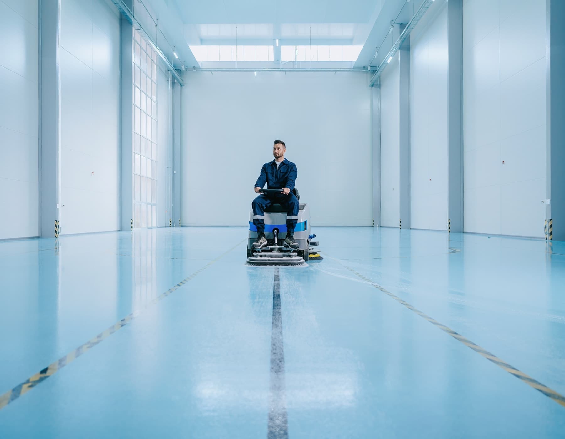 Medewerker bestuurt een opzitschrobmachine in een lege, lichte productiehal met epoxyvloer.